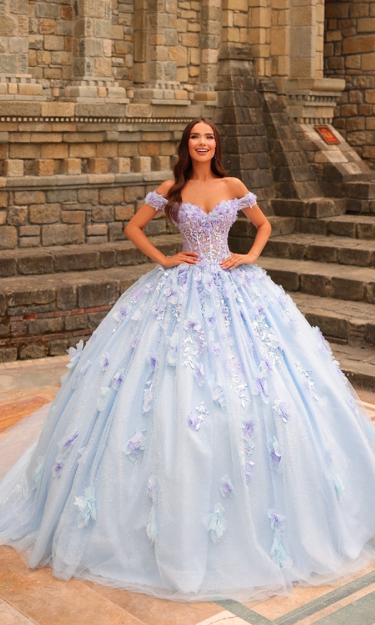 A woman stands outdoors by stone steps, wearing the Amarra 54231 3D Floral Quinceañera Dress—a light blue gown with off-the-shoulder sleeves and beautiful 3D floral decorations.